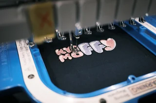 刺繍ミシンで、黒い生地に「KyaffyMoffy」のロゴをピンクと紫の糸で刺繍している様子